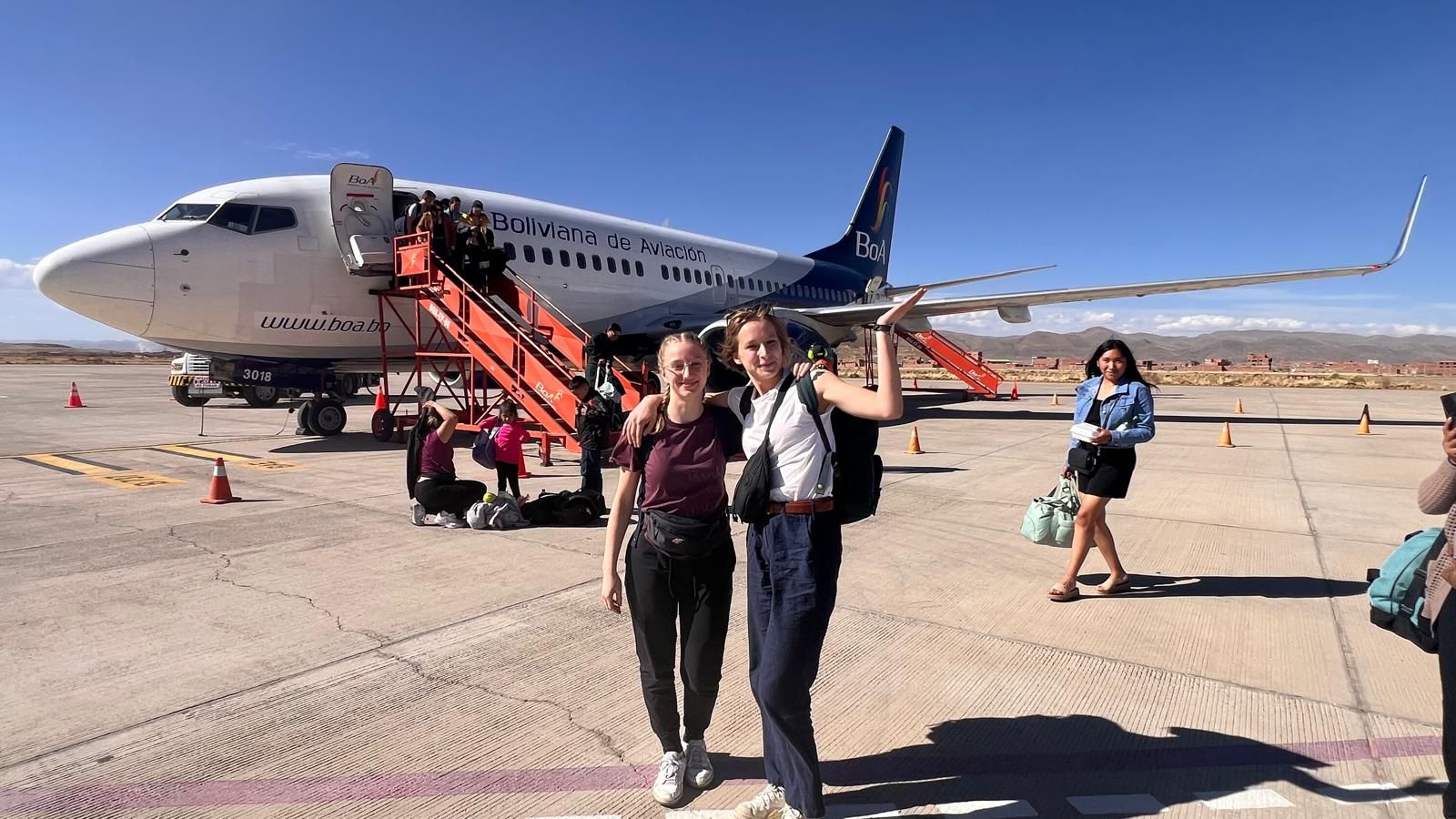 Na pierwszym planie Od lewej Weronika Tietz obok Zuzanna Krzywda na płycie lotniska Santa Cruz w tle samolot oraz pasażerowie.