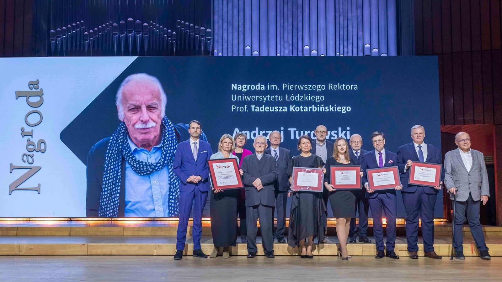 Grupowe zdjęcie laureatów oraz organizatorów Nagrody im. Pierwszego Rektora Uniwersytetu Łódzkiego, prof. Tadeusza Kotarbińskiego, na scenie z wielkim ekranem w tle. Na ekranie widnieje wizerunek Andrzeja Turczyńskiego, laureata nagrody, oraz nazwa wydarzenia.