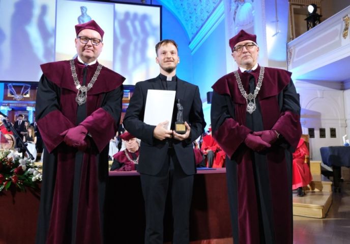 Na zdjęciu widzimy trzy osoby stojące na scenie podczas uroczystości akademickiej lub wręczenia nagród. Dwaj mężczyźni po bokach ubrani są w tradycyjne togi akademickie w kolorach bordowym i czarnym oraz nakrycia głowy w tym samym kolorze. Na piersiach mają łańcuchy rektorskie. Mężczyzna w środku, ubrany w czarny garnitur i koloratkę, trzyma w dłoniach dyplom oraz statuetkę. W tle widać inne osoby w togach. Na zdjęciu od lewej: prof. dr hab. Piotr Pawluć, ks. dr Przemysław Zgórecki, prof. UAM dr hab. Rafał Witkowski. Fot. Przemysław Stanula