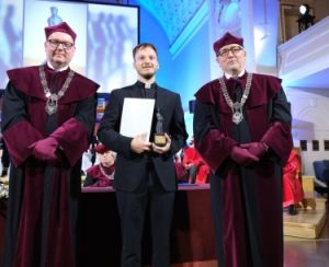 Na zdjęciu widzimy trzy osoby stojące na scenie podczas uroczystości akademickiej lub wręczenia nagród. Dwaj mężczyźni po bokach ubrani są w tradycyjne togi akademickie w kolorach bordowym i czarnym oraz nakrycia głowy w tym samym kolorze. Na piersiach mają łańcuchy rektorskie. Mężczyzna w środku, ubrany w czarny garnitur i koloratkę, trzyma w dłoniach dyplom oraz statuetkę. W tle widać inne osoby w togach. Na zdjęciu od lewej: prof. dr hab. Piotr Pawluć, ks. dr Przemysław Zgórecki, prof. UAM dr hab. Rafał Witkowski. Fot. Przemysław Stanula
