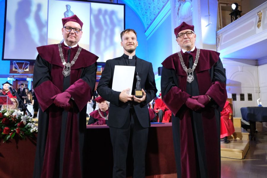 Na zdjęciu widzimy trzy osoby stojące na scenie podczas uroczystości akademickiej lub wręczenia nagród. Dwaj mężczyźni po bokach ubrani są w tradycyjne togi akademickie w kolorach bordowym i czarnym oraz nakrycia głowy w tym samym kolorze. Na piersiach mają łańcuchy rektorskie. Mężczyzna w środku, ubrany w czarny garnitur i koloratkę, trzyma w dłoniach dyplom oraz statuetkę. W tle widać inne osoby w togach. Na zdjęciu od lewej: prof. dr hab. Piotr Pawluć, ks. dr Przemysław Zgórecki, prof. UAM dr hab. Rafał Witkowski. Fot. Przemysław Stanula