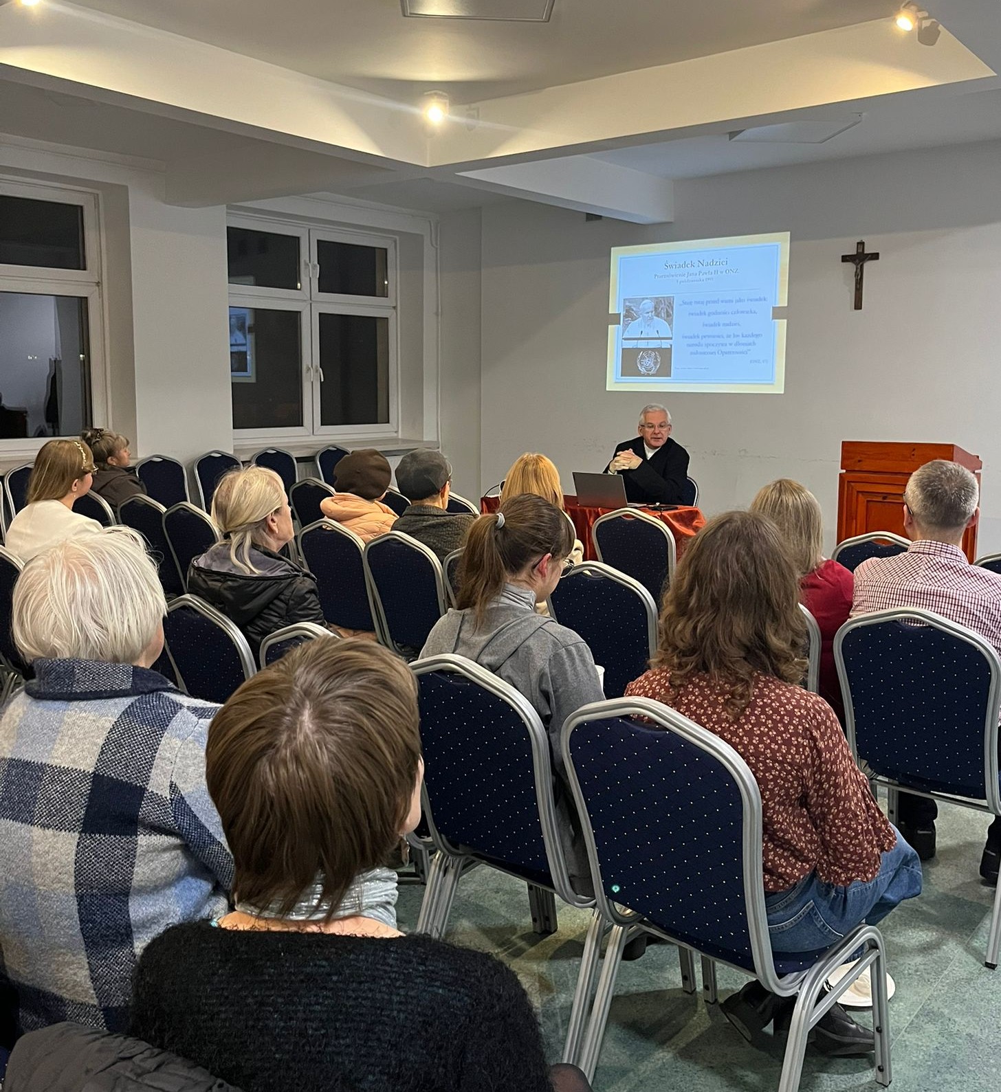 ks. prof. UAM dr hab. Maciej Olczyk siedząc przy stole przemawia, na pierwszym planie słuchacze wykładu otwartego