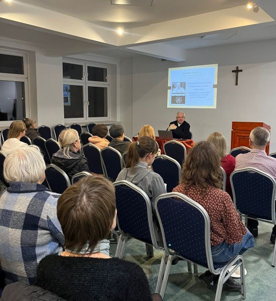 ks. prof. UAM dr hab. Maciej Olczyk siedząc przy stole przemawia, na pierwszym planie słuchacze wykładu otwartego